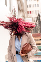 A woman with pink curly hair blowing in the wind. Standing on the stairs in a light raincoat and...