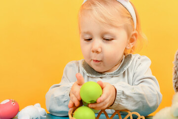 Fototapeta premium A little girl takes out painted Easter eggs from a basket and shows them to a toy bunny for Easter.