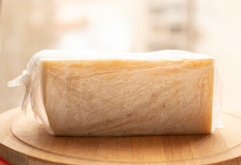 a piece of cheese in a plastic wrap lies on a wooden board, on a light background. Food, lactose-free cheese, healthy food, daily diet