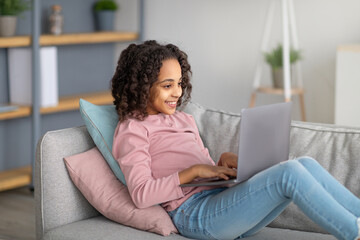 Fototapeta premium Youth and technology. Black girl using laptop and typing on keyboard, sitting on comfortable couch
