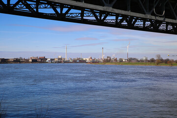 Fototapeta premium Krefeld (Uerdingen) - March 1. 2021: View beyond steel bridge over river rhine on industrial area with factories and chimneys against blue sky