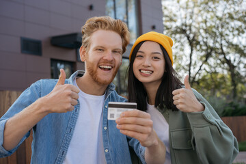 Couple of smiling mixed race friends holding credit card showing thumb up looking at camera