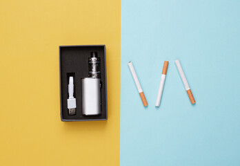 Vaping device in box and cigarettes on yellow blue background. Alternative to smoking. Top view
