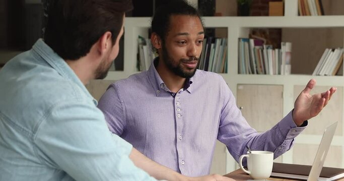 Diverse multi racial workmates discuss collaborative task in office. African and Caucasian male colleagues talking sit at workplace desk share ideas, skills, develop strategy solve business at meeting