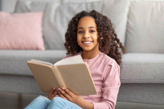 Bookworm. Cheerful black teen girl reading paper book, resting at home and smiling to camera