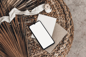 Tropical summer mockups. Black cell phone with empty screen, blank greeting card, envelope and ribbon placed on rattan straw pouff. Dry palm leaves. Minimal modern boho interior, flat lay, top view. © tabitazn