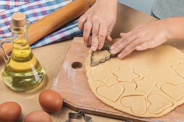 The child's hands hold a metal cookie cutter and cut homemade cookies of various shapes from shortcrust pastry. On table rolling pin, eggs, sunflower oil, towel.  Close up, top and side view.