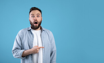 Unbelievable news. Young bearded guy opening mouth in shock, pointing at blank space over blue...