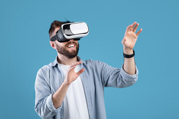 Naklejka premium Happy bearded young man using VR headset to explore augmented reality on blue studio background