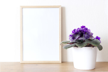 Horizontal white frame mockup on vintage wooden table. Modern white ceramic vase with Violet Saintpaulias flowers White wall background. Selective focus.