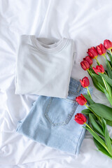 Female trendy clothes flat lay on white background, women minimalistic set of blue jeans and gray sweatshirt with bouquet of red tulips top view