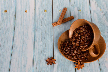 The cup lies on its side on a saucer with coffee beans in it. Next to it lies anise and cinnamon on a wooden background