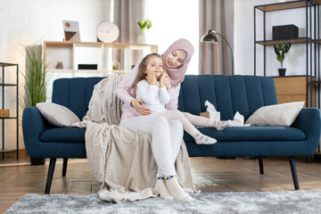 Obraz premium Happy Arabian mother sits on sofa at home and hugs her little daughter. Adorable smiling little girl after bath, with cream on her face, copying mom behavior, using cosmetic cream at home