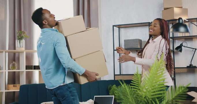 Attractive satisfied young black-skinned man carrying carton boxes into his new house where beautiful stylish wife is waiting for him to help,family relocation concept