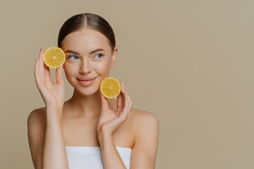 Horizontal shot of thoughtful young European woman uses homemade fruit for facial mask holds lemon slices wrapped in white soft bath towel looks aside isolated over brown background copy space