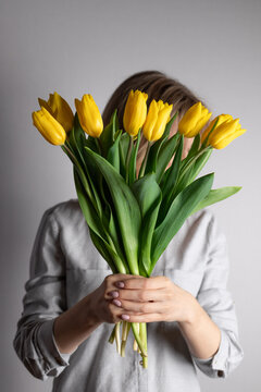 Woman holding bouquet of yellow tulips and hiding behind it against gray wall. Floral lifestyle composition. vertical image.