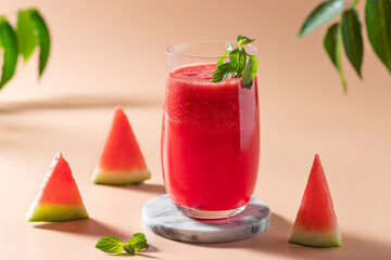 glass of freshly squeezed watermelon juice with mint, watermelon slices and green leaves in background. Colorful refreshing drinks for summer. beige background. space for text