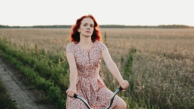 Portrait of dreamy romantic girl in sundress on vintage retro bicycle at meadow in sunset light. Woman is cycling on old bike through summer road. Happy Beautiful Girl Relaxing. Happy lifestyle.