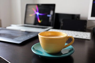 yellow coffee cup with green saucer on desk with computers, telework concept.