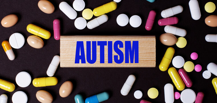 On a dark background, multi-colored pills and the word AUTISM on a wooden block. Medical concept