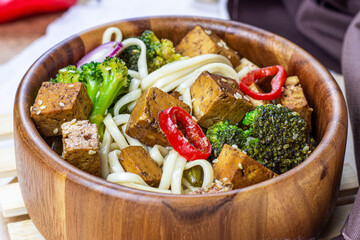 Homemade vegetarian fried tofu with udon noodles and vegetables (broccoli, pepper and onions) in the wooden bowl on light kitchen background.