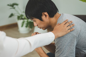 Young asian man suffers from a mental who needs to therapy with a psychologist while sitting on couch to consult, psychiatrist has encouragement the patient by touching to make his feel relaxed.