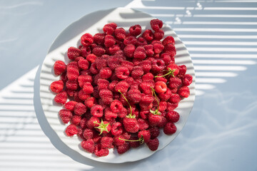 Natural ripe red raspberries on a white plate. Shade from the blinds. Selective focus.