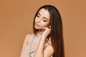 Portrait of the young woman with perfect trendy makeup in an elegant beige dress posing in studio on the beige background. Concept of modish evening makeup and smooth skin