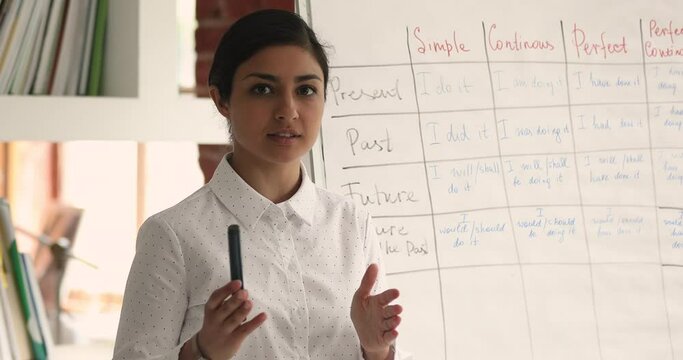 Indian mixed race teacher standing near flip chart in classroom looking at camera telling about foreign language tenses, teaches English grammar using video call. Remote education and tutoring concept
