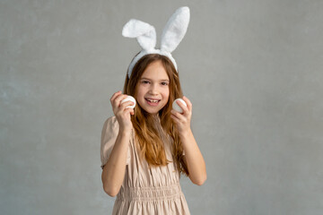 Funny kid celebrating Christian holiday. Closeup portrait of girl. grey background with white eggs....