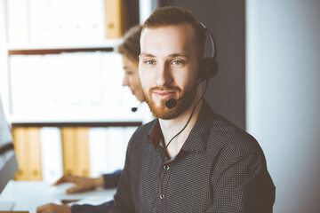 Red-bearded businessman talking by headset near his female colleague while sitting in sunny office. Diverse people group in call center