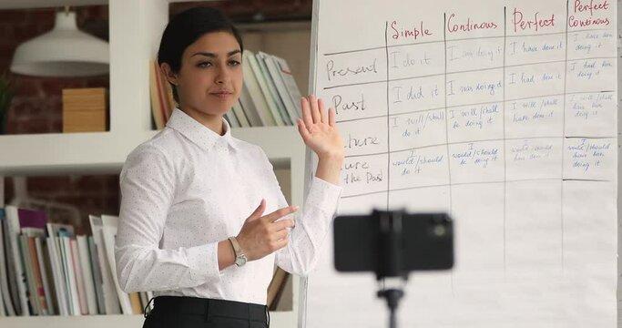 Indian female young teacher blogger standing in front of smart phone on tripod record education English grammar class, explain tenses on flip chart. Improve language skill, tutoring, education concept