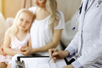 Fototapeta premium Doctor and patient. Pediatrician using clipboard while examining little girl with her mother at home. Happy cute caucasian child at medical exam. Medicine concept