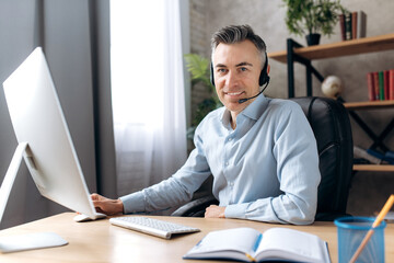 Portrait of successful confident adult caucasian male in formal wear, businessman or lawyer sitting at workplace in modern office using computer and headset, looking at camera with friendly smile