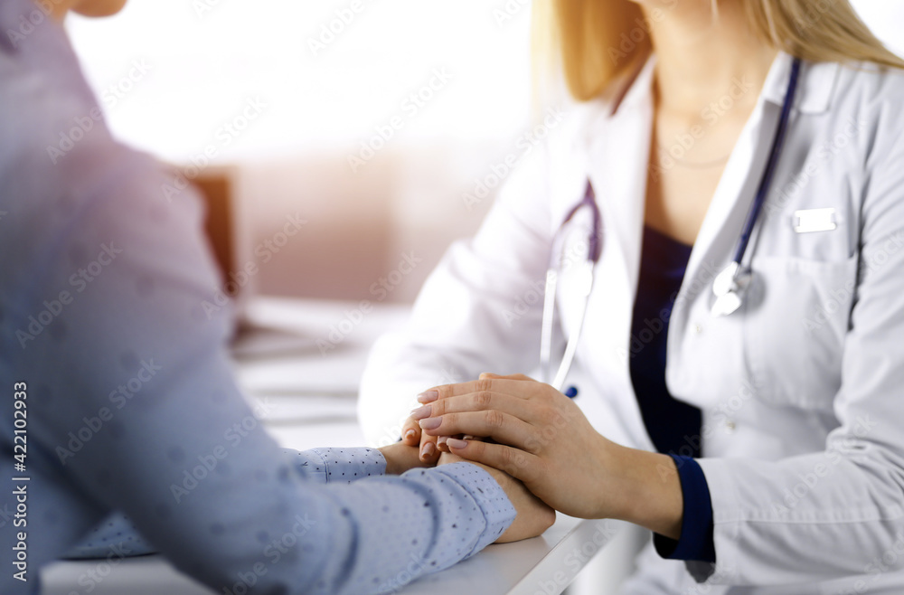 Wall mural Unknown woman-doctor is holding her patient's hands to reassure the patient, discussing current health examination, while they are sitting together at the desk in the sunny cabinet in a clinic. Female