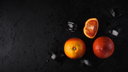 ripe red whole and cut oranges with ice cubes and water drops lie on the side on a black textured plaster. top view. artistic moody food photo with copy space