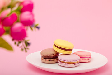 Plate of macaroons on pink background with flowers. Sweet pastry, baked products, sweets, dessert. Unhealthy diet, sugar dependence.