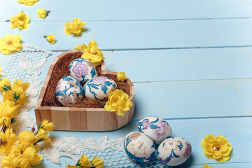 Decorated Easter eggs in wooden heart shaped box. White lace, yellow artificial flowers on blue background. Place for text.