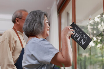 Happy retired senior Asian couple standing in the house as a small business restaurant putting opening sign on the door with happy welcoming smiley face