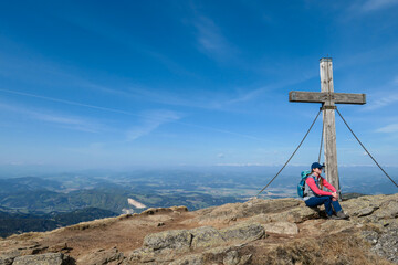 A woman with hiking backpack sitting under a wooden cross on top of Sauofen in Austrian Alps. A vast, golden pasture around. Fall vibes. Mountain chains in the back. Panoramic view. Achievement