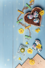 Heart shaped box with decorated Easter eggs, yellow flowers, burning candle and brown boards on wooden blue background. Spring and Easter concept. Top view, flatlay, copy space. Vertical shot.