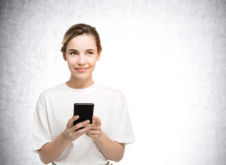 Portrait of attractive young woman holding smartphone smiling and dreaming about pleasant things. Concrete wall on background.