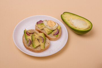 Two sandwiches with avocado, boiled eggs and red onions on white plate, avocado half on beige background. Minimalistic style composition.