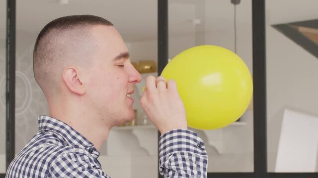 close up in profile young white dark-haired man smiles and inflates and blows the yellow ballon at home