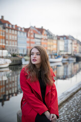 Fototapeta premium Attractive girl sitting, she is a tourist and is visiting Nyhavn in Copenhagen, Denmark. She is wearing a red coat