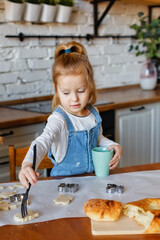 The girl is standing at the table in the kitchen playing with dough and cookie cutters.