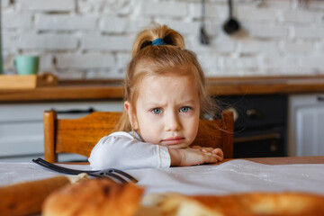 Portrait of a little girl 3 years old sitting at the kitchen table. A sad child is having breakfast in the kitchen.