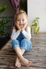 A little blonde girl with blue eyes in a denim jumpsuit is sitting on a large wooden window sill near the window.