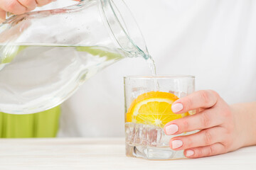 Female hands pouring water from the decanter into a glass beaker with lemon and ice. Health and...