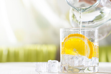 Female hands pouring water from the decanter into a glass beaker with lemon and ice. Health and...